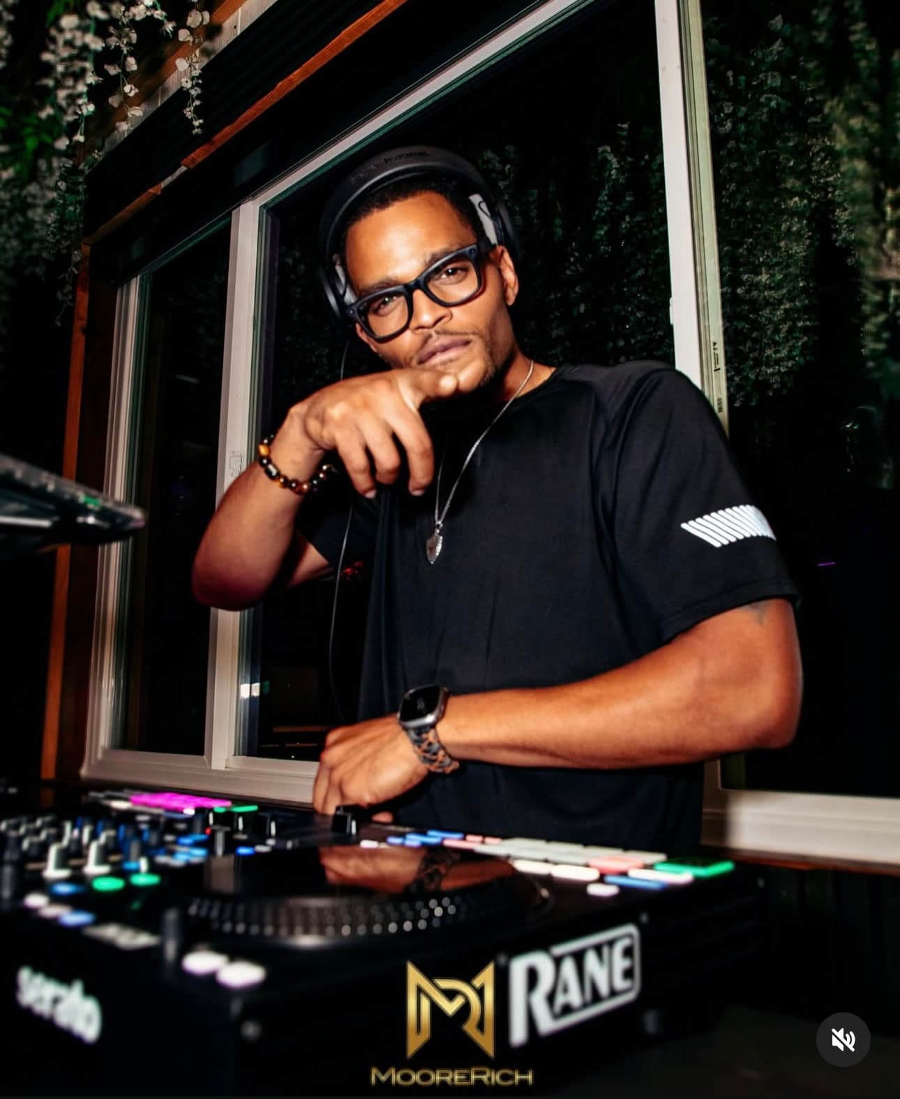DJ Waterzz standing beside a mixing controller in a dim room.